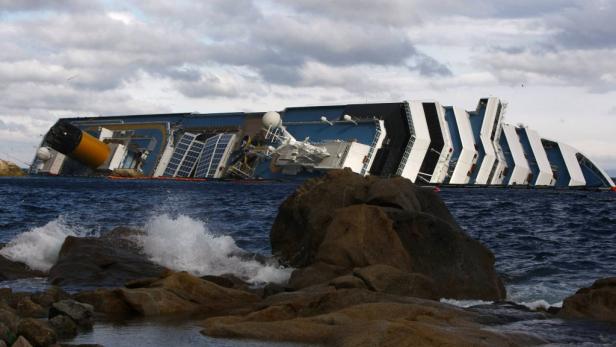 Ein großes Kreuzfahrtschiff liegt auf der Seite im Meer, nahe felsiger Küste.