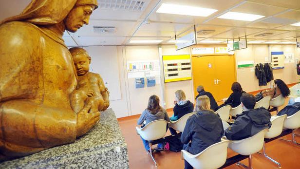 Menschen warten in einem Wartezimmer vor einer Statue von Maria mit Kind.