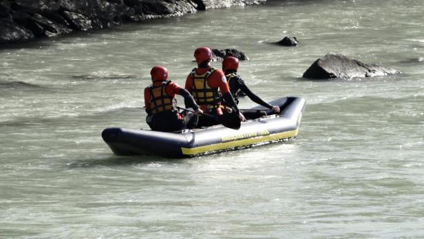 Drei Personen in Neoprenanzügen und Helmen sitzen in einem Schlauchboot auf einem Fluss.