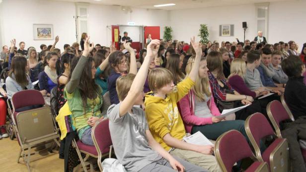 Viele junge Leute sitzen in einem Raum und heben ihre Hände.