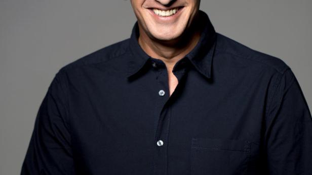 Ein lächelnder Mann mit dunklem Haar und blauem Hemd vor grauem Hintergrund.