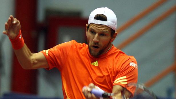 Ein Tennisspieler in einem orangefarbenen Trikot und einer weißen Kappe schlägt einen Ball.