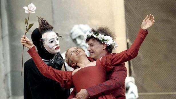 Eine Szene mit drei Darstellern, einer mit Maske und Rose, ein Paar in roter Kleidung, vor einer Mauer.