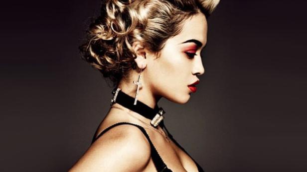 Eine Frau mit blonden Locken, rotem Lidschatten und Halsband im Profil vor dunklem Hintergrund.