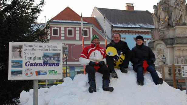 Drei Personen sitzen auf einem Schneehaufen vor einem Werbeschild für das Langlaufzentrum Gutenbrunn.