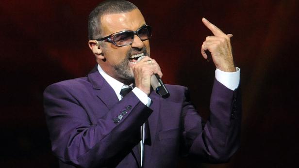 George Michael singt in einem lila Anzug auf der Bühne und zeigt mit dem Finger nach oben.