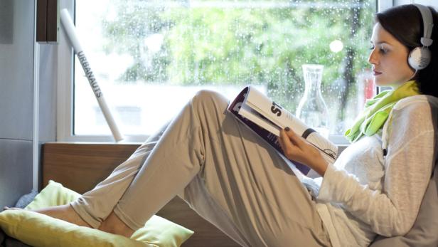Eine Frau sitzt am Fenster, liest eine Zeitschrift und hört Musik über Kopfhörer.