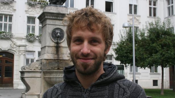 Ein Mann mit Bart und lockigem Haar steht vor einem Brunnen und einem Gebäude.