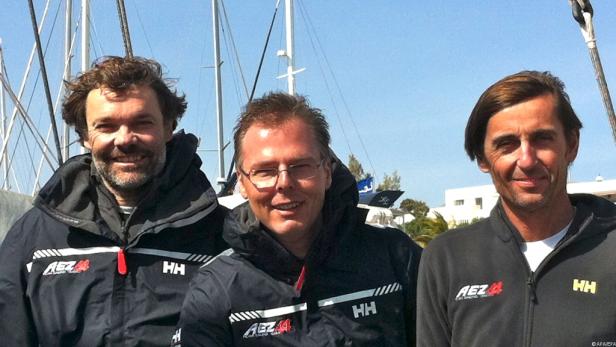 Drei Männer in Segeljacken posieren vor einem Hintergrund mit Segelbooten.
