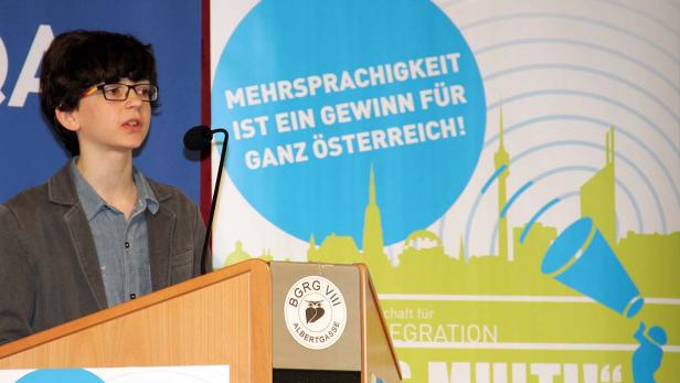 Ein junger Redner steht an einem Pult mit dem Slogan „Mehrsprachigkeit ist ein Gewinn für ganz Österreich!“.