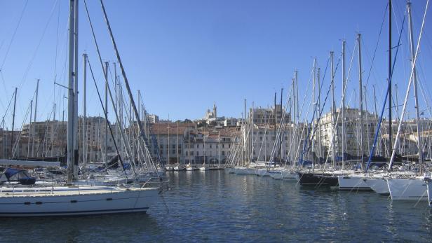 Blick auf den Hafen von Marseille mit zahlreichen Segelbooten und der Basilika Notre-Dame de la Garde im Hintergrund.
