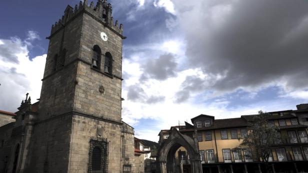 Ein hoher Kirchturm aus Stein steht auf einem Platz unter bewölktem Himmel.