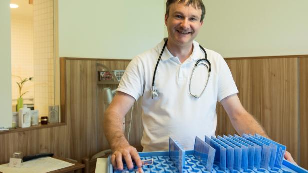 Ein lächelnder Arzt mit Stethoskop steht vor einem blauen Tablett mit Medikamenten.