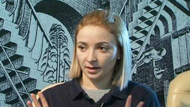 Eine Frau mit blonden Haaren gestikuliert vor einem Wandbild mit einer Leiter und einer Person.