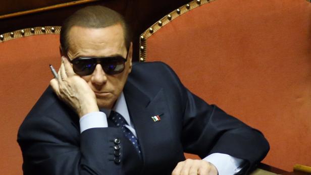 Silvio Berlusconi mit Sonnenbrille und Stift in der Hand sitzt auf einem Stuhl.