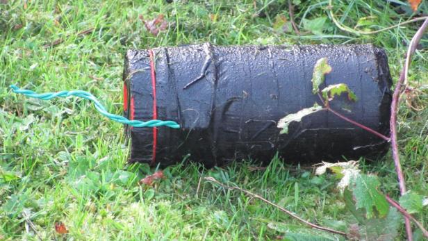 Ein zylinderförmiger Gegenstand, umwickelt mit schwarzem Klebeband und grünen Drähten, liegt im Gras.