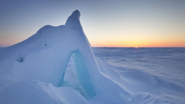 Ein Eisberg mit einer bogenförmigen Öffnung in einer verschneiten Landschaft bei Sonnenuntergang.