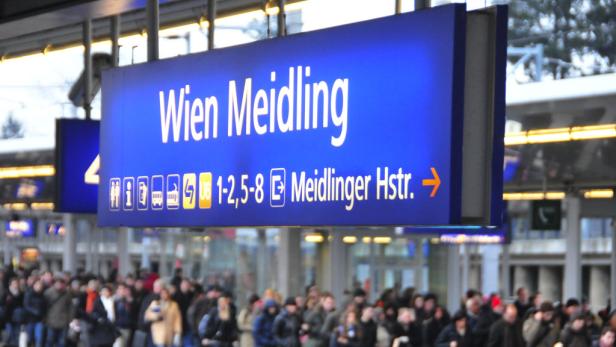 Ein blaues Schild mit der Aufschrift „Wien Meidling“ hängt in einem Bahnhof über einer Menschenmenge.