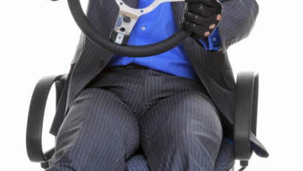 Ein Mann im Anzug und mit Fliegerbrille sitzt auf einem Bürostuhl und hält ein Lenkrad.