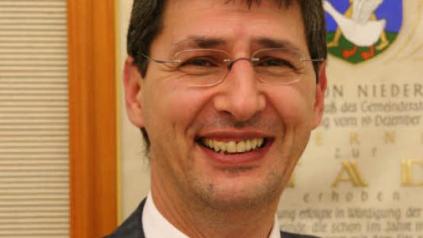 Ein lächelnder Mann mit Brille und Anzug vor einem Wappen.