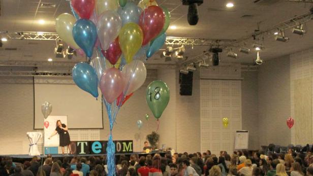 Eine Gruppe von Menschen sitzt auf dem Boden vor einer Bühne mit Ballons.
