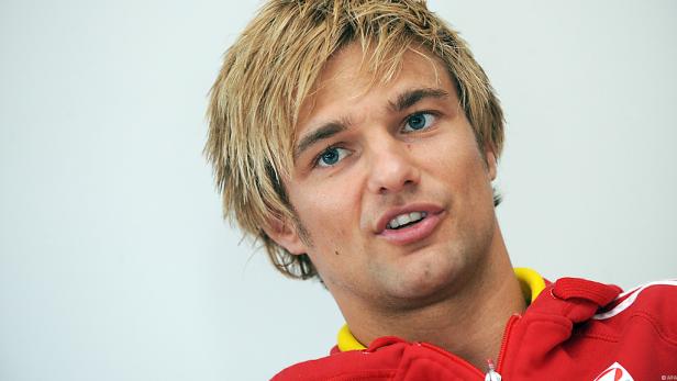 Ein Mann mit blonden Haaren und blauen Augen in einem roten Trainingsanzug.