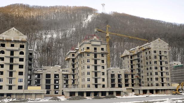 Mehrere Rohbauten stehen vor einem bewaldeten Hügel mit einem Baukran.