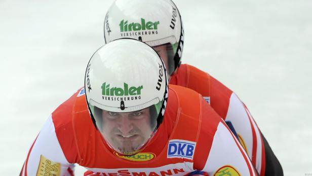 Zwei Rennrodler mit Helmen und roter Sportbekleidung während eines Wettkampfs.