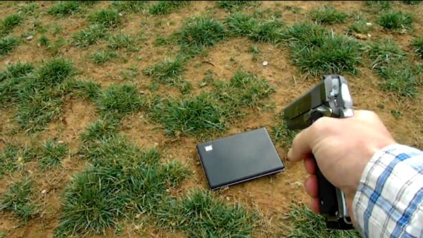Eine Person zielt mit einer Pistole auf einen Laptop, der auf dem Boden liegt.