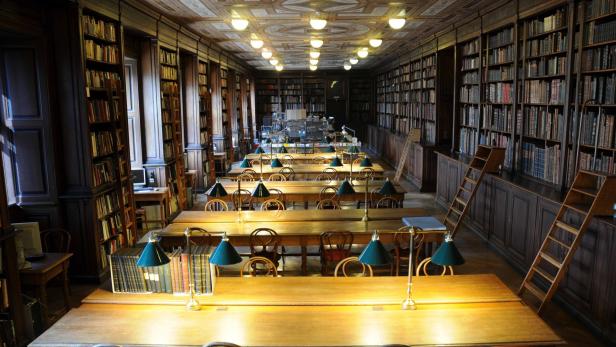 Ein langer Lesesaal mit Holztischen, grünen Lampen und hohen Bücherregalen.