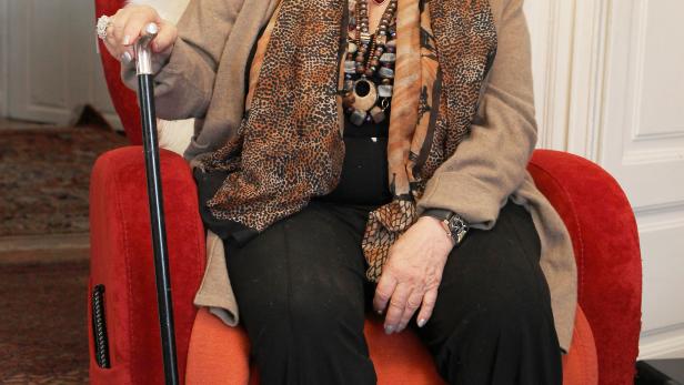 Eine ältere Frau mit rotem Haar sitzt mit einem Gehstock in einem Sessel.