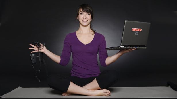 Eine Frau sitzt im Schneidersitz auf einer Yogamatte und hält einen Laptop und Kopfhörer.