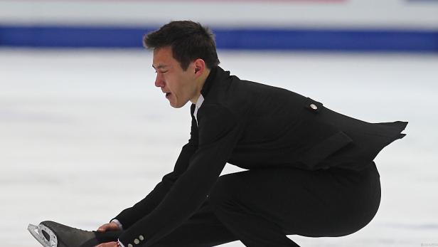 Ein Eiskunstläufer in schwarzer Kleidung sitzt auf dem Eis und schnürt seine Schlittschuhe.