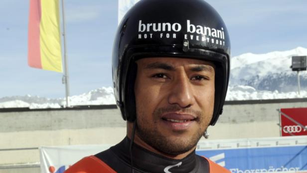 Ein Rennrodler mit Helm der Marke Bruno Banani vor einer verschneiten Bergkulisse.