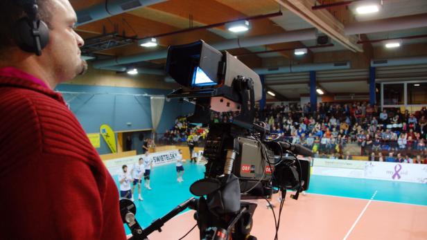 Ein Kameramann filmt ein Volleyballspiel in einer Halle mit Zuschauern.