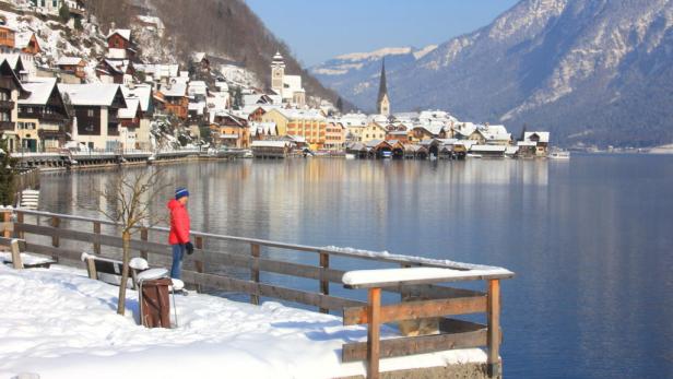 Eine Person in roter Jacke steht an einem verschneiten Ufer mit Blick auf Hallstatt.