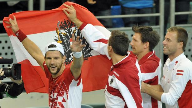 Ein Tennisspieler jubelt mit der österreichischen Flagge im Arm, umgeben von seinem Team.