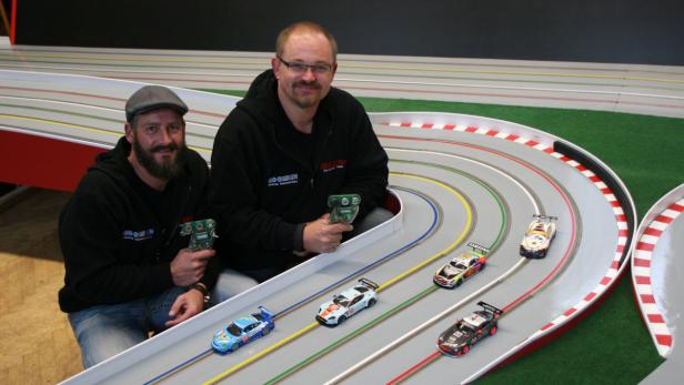 Zwei Männer sitzen an einer Autorennbahn und halten die Controller in der Hand.
