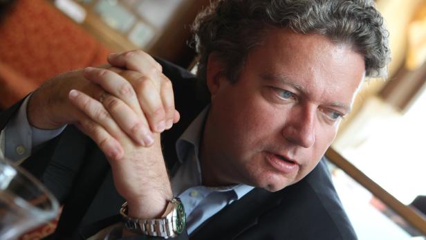 Ein Mann mit grauen Haaren und Anzug sitzt mit verschränkten Händen da.