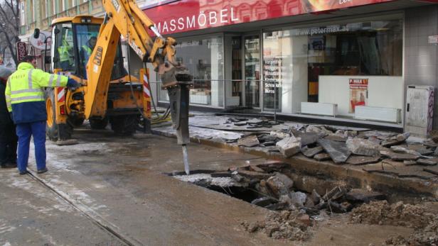 Ein Bagger mit Presslufthammer reißt vor einem Möbelgeschäft den Asphalt auf.