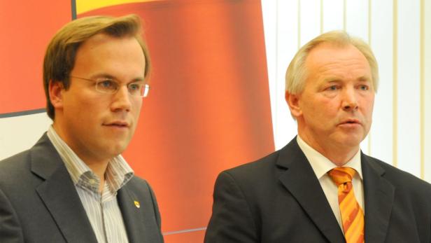 Zwei Männer in Anzügen stehen vor einem roten Hintergrund.