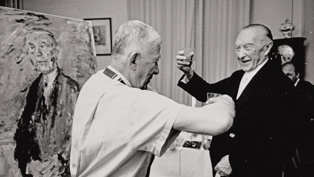 Ein Mann mit Orden prostet einem älteren Mann mit Anzug und Glas zu, im Hintergrund ein Porträtgemälde.