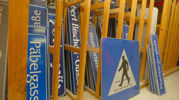 Ein Lagerregal voller blauer Straßenschilder, darunter „Päbelgasse“ und ein Fußgängerüberweg.
