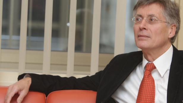 Ein Mann mit Brille und Anzug sitzt auf einem orangefarbenen Sofa.