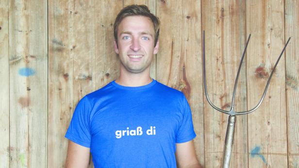 Ein Mann mit blauem T-Shirt und Heugabel vor einer Holzwand.