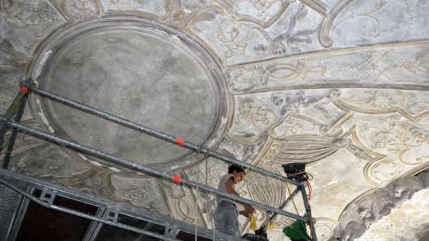Eine Restauratorin arbeitet auf einem Gerüst an der Stuckdecke eines alten Gebäudes.