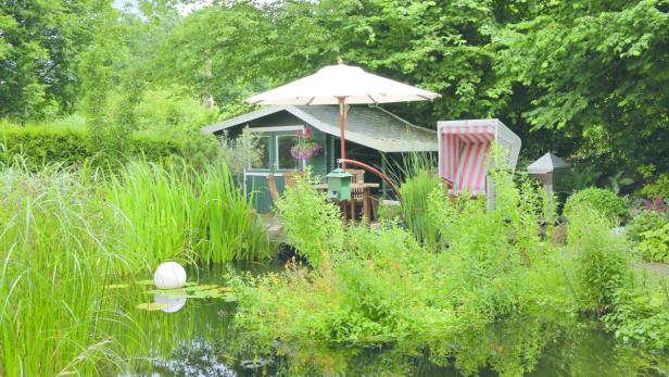 Ein Garten mit einem Teich, einem kleinen Häuschen und einem Strandkorb.