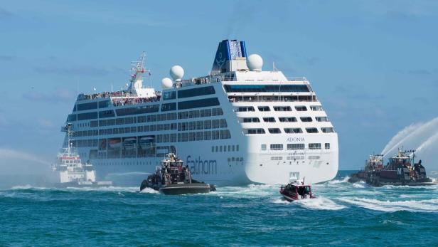 Das Kreuzfahrtschiff „Adonia“ wird von Schleppern mit Wasserfontänen auf dem Meer begleitet.