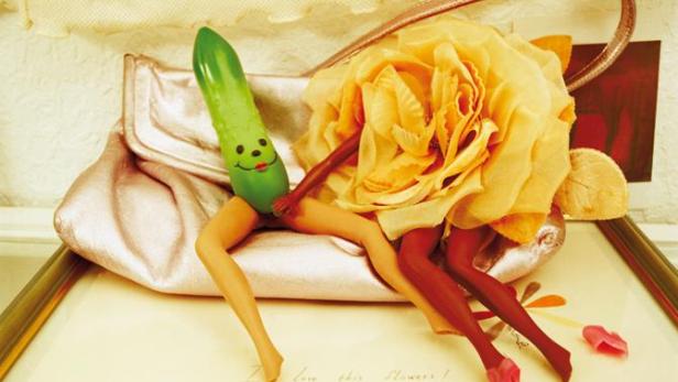 Eine Gurke mit Gesicht und zwei Paar Beine posieren auf einer Handtasche und einer gelben Rose.