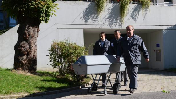 Drei Männer schieben einen grauen Sarg auf einem Rollwagen.